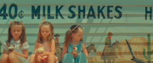 frame from the film Asteroid City. three small girls lean against the side of a painted building, each holding a milkshake.