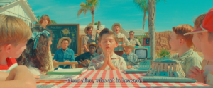 frame from the film Asteroid City. a small boy holds his hands in prayer while singing the opening bars to the film's musical number "Dear Alien (Who Art In Heaven)".